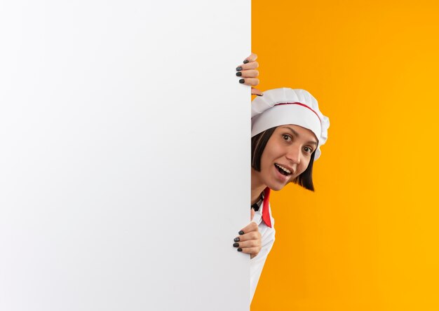 Gioiosa giovane donna cuoca in uniforme da chef guardando la parte anteriore da dietro il muro bianco isolato sull'arancio