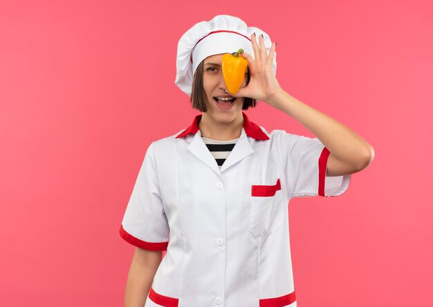 Gioiosa giovane donna cuoca in uniforme da chef guardando la parte anteriore da dietro pepe isolato sul colore rosa