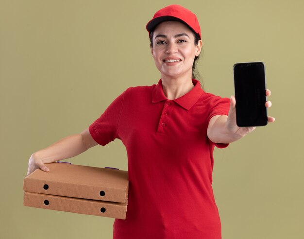 Gioiosa giovane donna delle consegne in uniforme e berretto che tiene in mano i pacchetti di pizza che allungano il telefono cellulare