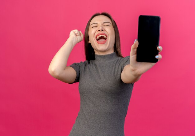 Gioiosa giovane donna graziosa che allunga il telefono cellulare alla telecamera alzando il pugno con gli occhi chiusi isolato su sfondo rosa con spazio di copia