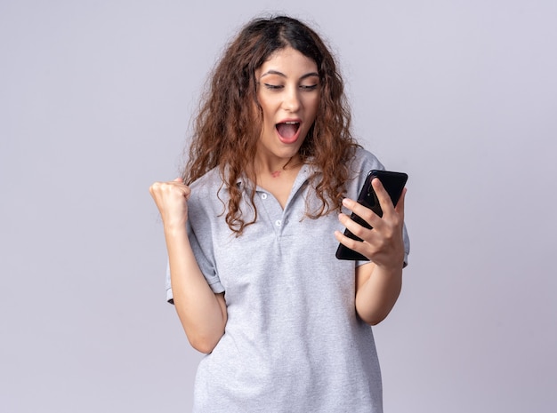 Gioiosa giovane donna graziosa che tiene e guarda il telefono cellulare facendo sì gesto isolato sul muro bianco con copia spazio