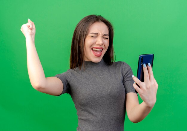 Gioiosa giovane donna graziosa che tiene il telefono cellulare alzando il pugno facendo sì gesto con gli occhi chiusi isolati su sfondo verde