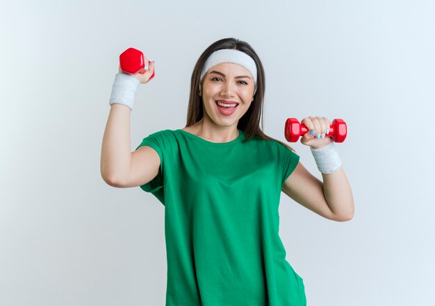 Gioiosa giovane donna sportiva che indossa la fascia e braccialetti cercando di alzare i manubri