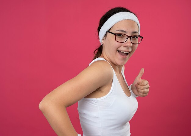 Gioiosa giovane donna sportiva in occhiali ottici che indossa la fascia e braccialetti si leva in piedi lateralmente e pollice in alto isolato sulla parete rosa