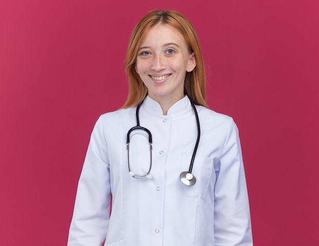 Gioiosa giovane dottoressa allo zenzero che indossa un abito medico e uno stetoscopio sorridente