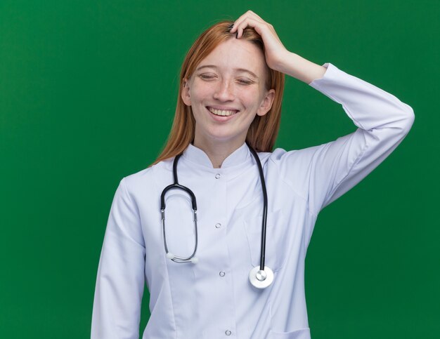 Gioiosa giovane dottoressa allo zenzero che indossa una tunica medica e uno stetoscopio che tocca la testa sorridendo con gli occhi chiusi isolati sul muro verde