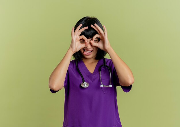 Gioiosa giovane dottoressa bruna in uniforme con lo stetoscopio sporge la lingua guardando attraverso le dita isolate su sfondo verde oliva con spazio di copia