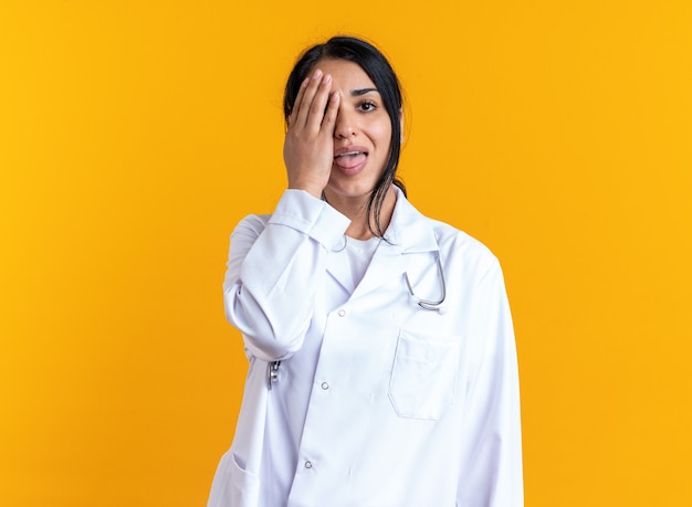 Gioiosa giovane dottoressa che indossa un abito medico con uno stetoscopio coperto l'occhio con la mano isolata sulla parete gialla