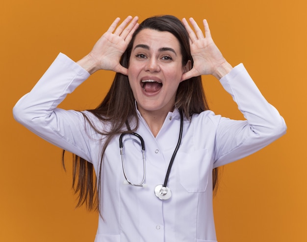 Gioiosa giovane dottoressa che indossa un abito medico con uno stetoscopio si tiene per mano vicino alla testa isolata sulla parete arancione con spazio di copia