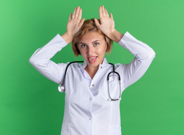 Gioiosa giovane dottoressa che indossa una veste medica con uno stetoscopio che mostra il gesto delle orecchie da coniglio isolato sulla parete verde green