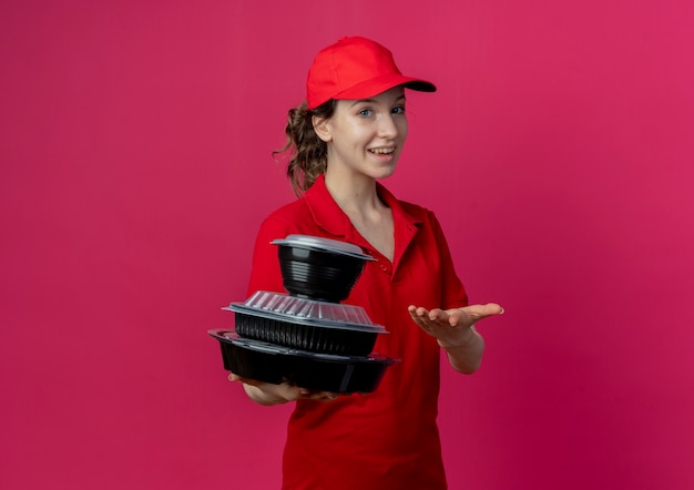 Gioiosa giovane graziosa ragazza delle consegne che indossa l'uniforme rossa e il berretto che tiene e indica con la mano i contenitori per alimenti