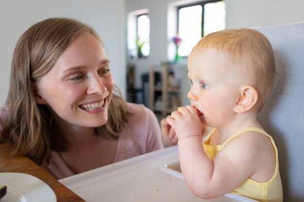 Gioiosa giovane mamma guardando la figlia del bambino che morde il pezzo di cibo. Colpo del primo piano. La cura dei bambini o il concetto di nutrizione