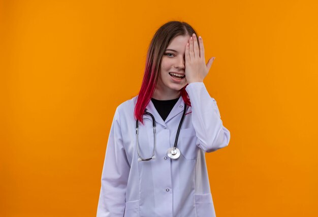 Gioiosa giovane medico donna che indossa stetoscopio abito medico coperto occhio con la mano sulla parete arancione isolata