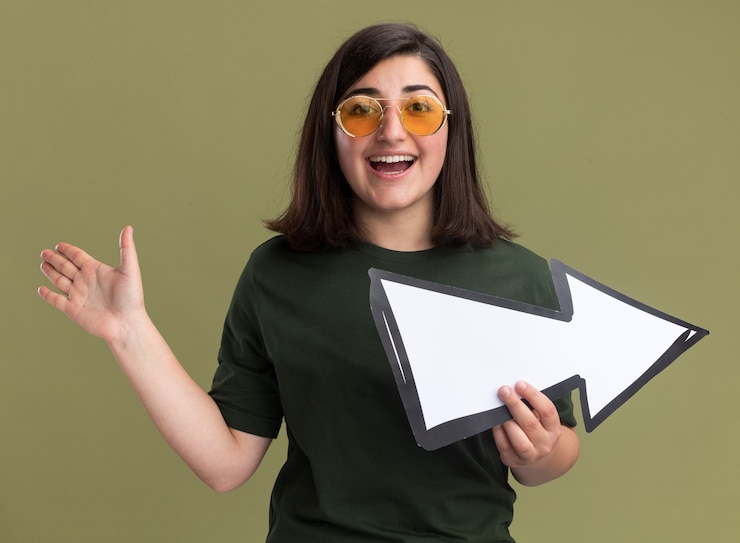 Foto gratuito gioiosa giovane ragazza abbastanza caucasica in occhiali da sole sta con la mano alzata e tiene il segno della freccia su verde oliva