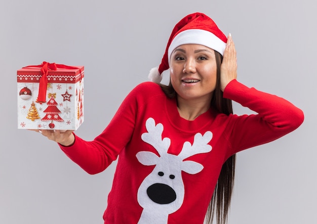 Gioiosa giovane ragazza asiatica che indossa il cappello di Natale con il maglione tenendo il contenitore di regalo mettendo la mano sulla guancia isolati su sfondo bianco