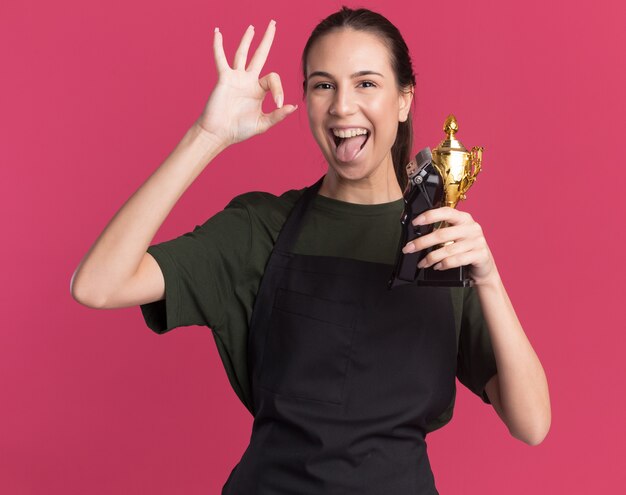 Gioiosa giovane ragazza bruna barbiere in uniforme sporge la lingua gesticolando segno giusto della mano che tiene i tagliacapelli e la tazza del vincitore sul rosa