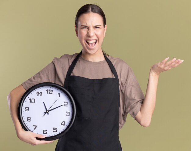 Gioiosa giovane ragazza bruna barbiere in uniforme tiene la mano aperta e tiene l'orologio su verde oliva