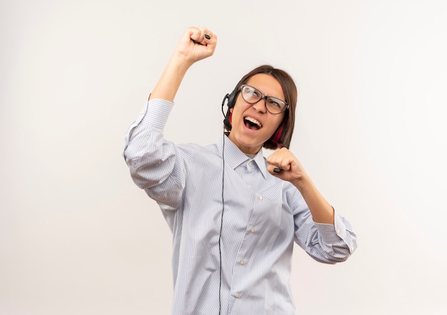 Gioiosa giovane ragazza call center con gli occhiali e auricolare innalzamento e guardando il pugno isolato su bianco