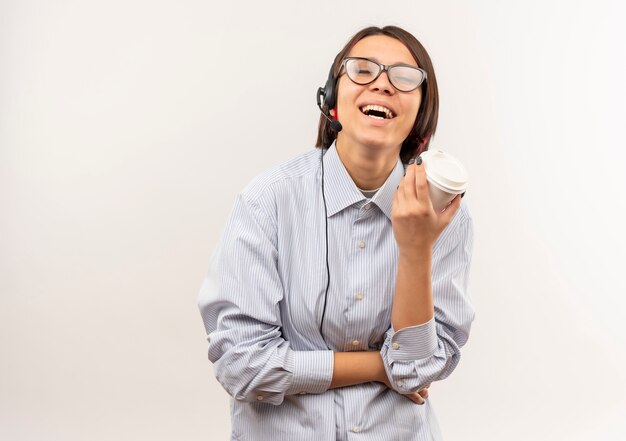 Gioiosa giovane ragazza call center con gli occhiali e auricolare tenendo la tazza di caffè in plastica con gli occhi chiusi isolati su bianco