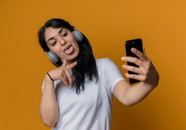 Gioiosa giovane ragazza caucasica bruna in cuffia guarda e punta al telefono isolato sulla parete arancione