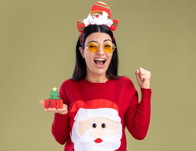 Gioiosa giovane ragazza caucasica che indossa la fascia di Babbo Natale e maglione con gli occhiali che tiene il giocattolo dell'albero di Natale con data ammiccante che fa gesto di sì isolato sulla parete verde oliva