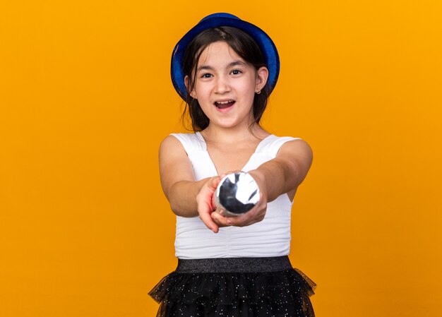 gioiosa giovane ragazza caucasica con cappello blu del partito che tiene il cannone dei coriandoli isolato sulla parete arancione con lo spazio della copia