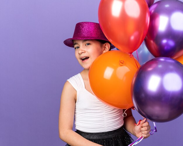 gioiosa giovane ragazza caucasica con cappello da festa viola che tiene palloncini di elio isolati su parete viola con spazio di copia