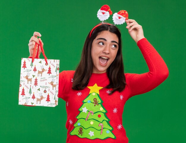 Gioiosa giovane ragazza caucasica con fascia santa tiene il sacchetto regalo di carta guardando il lato isolato su sfondo verde con spazio di copia