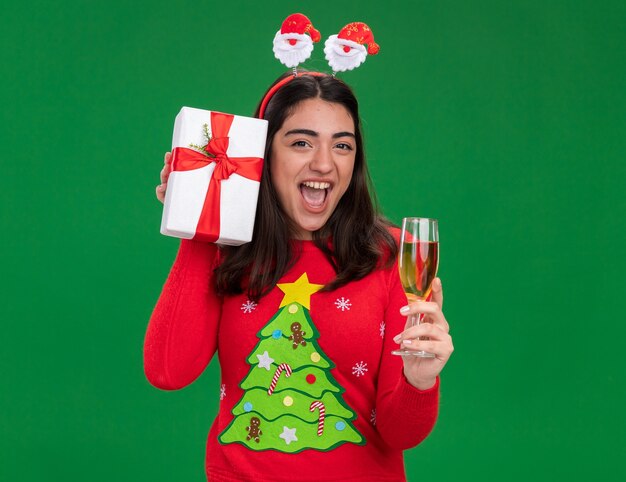 gioiosa giovane ragazza caucasica con fascia santa tiene un bicchiere di champagne e confezione regalo di Natale isolato su sfondo verde con spazio di copia