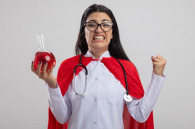 Gioiosa giovane ragazza caucasica del supereroe con gli occhiali e lo stetoscopio che tiene la boccetta chimica con liquido rosso facendo sì gesto che guarda l'obbiettivo isolato su priorità bassa bianca