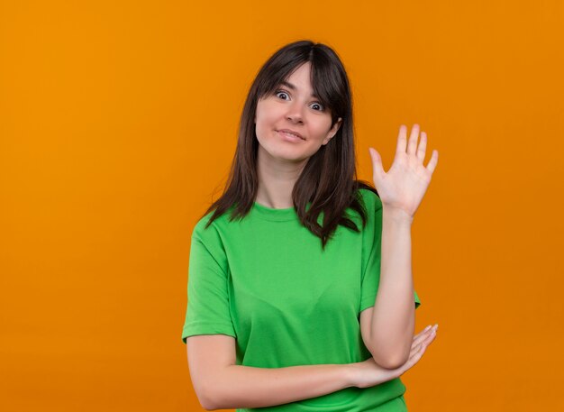 Gioiosa giovane ragazza caucasica in camicia verde alza la mano e guarda la telecamera su sfondo arancione isolato con spazio di copia