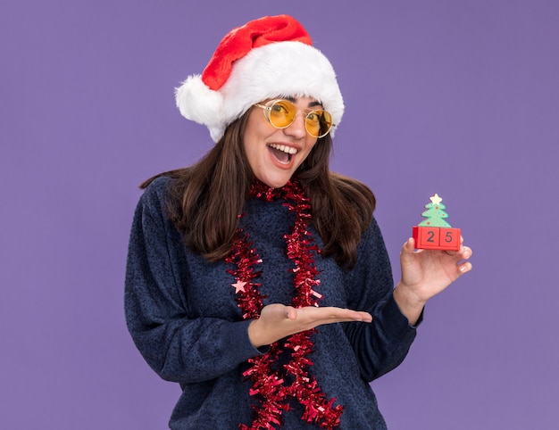 Gioiosa giovane ragazza caucasica in occhiali da sole con cappello da Babbo Natale e ghirlanda intorno al collo tiene e punta all'ornamento dell'albero di Natale isolato sulla parete viola con spazio di copia