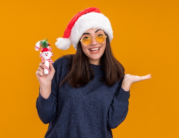 Gioiosa giovane ragazza caucasica in occhiali da sole con cappello da Babbo Natale tiene un bastoncino di zucchero e tiene la mano aperta isolata sul muro arancione con spazio per le copie