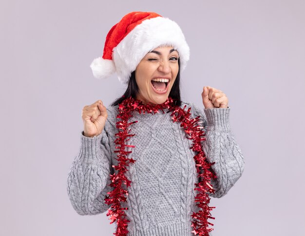 Gioiosa giovane ragazza caucasica indossando il cappello di Natale e la ghirlanda di orpelli intorno al collo che guarda l'obbiettivo ammiccante facendo sì gesto isolato su priorità bassa bianca