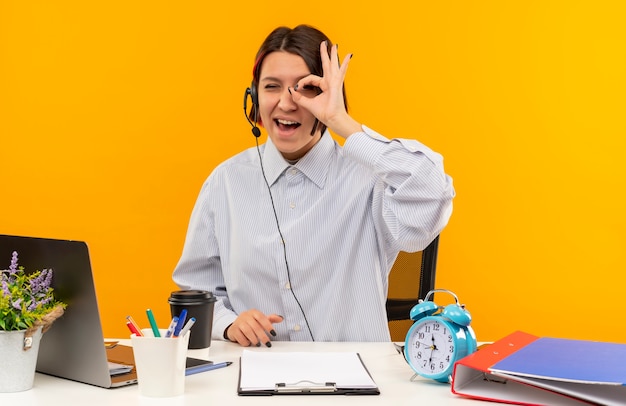 Gioiosa giovane ragazza della call center che indossa la cuffia avricolare che si siede allo scrittorio che fa il gesto di sguardo sbattere le palpebre isolato sull'arancio