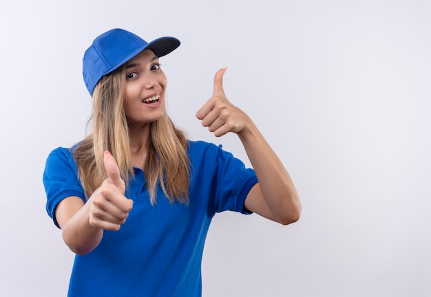 Gioiosa giovane ragazza di consegna che indossa l'uniforme blu e berretto i pollici in su isolato sulla parete bianca con lo spazio della copia