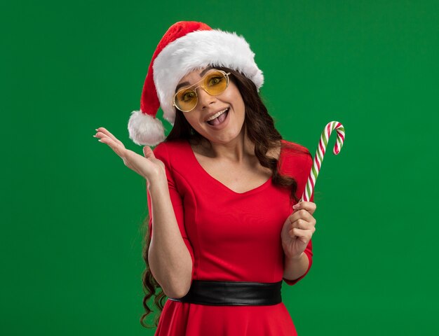 Gioiosa giovane ragazza graziosa che indossa il cappello della santa e bicchieri tenendo il bastoncino di zucchero di Natale guardando la telecamera mostrando la mano vuota isolata su sfondo verde