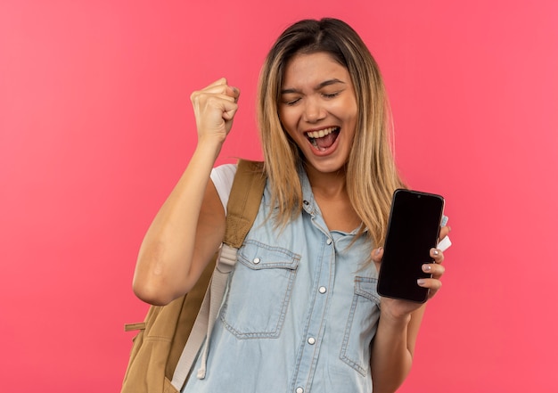 Gioiosa giovane ragazza graziosa dell'allievo che porta la borsa posteriore che tiene il telefono cellulare che alza il pugno con gli occhi chiusi isolati sul colore rosa