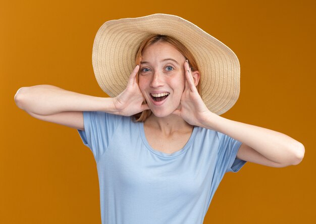 Gioiosa giovane ragazza rossa allo zenzero con le lentiggini che indossa un cappello da spiaggia mette le mani sul viso isolato sulla parete arancione con spazio di copia