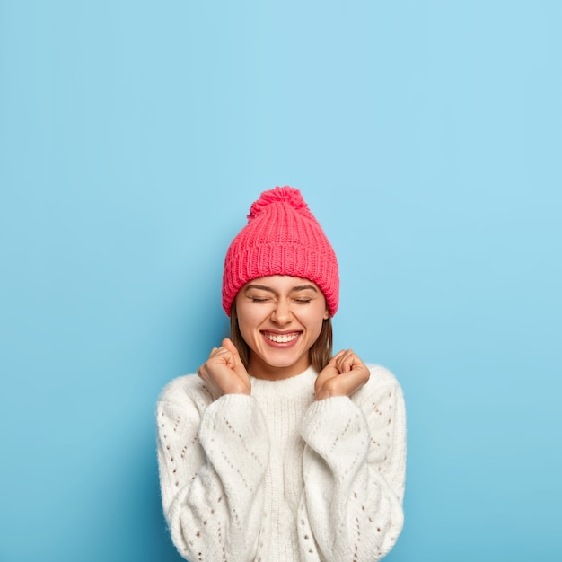 Gioiosa giovane ragazza si sente felicissima, solleva i pugni chiusi, è di buon umore, indossa un maglione bianco e cappello rosa, vestita con abiti caldi durante la fredda giornata autunnale, isolato sul muro blu