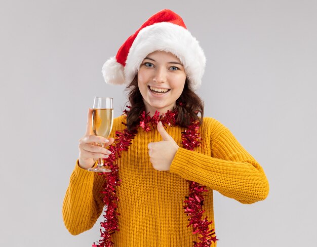 Gioiosa giovane ragazza slava con cappello da Babbo Natale e con ghirlanda intorno al collo tenendo un bicchiere di champagne e pollice in alto isolato sul muro bianco con spazio copia