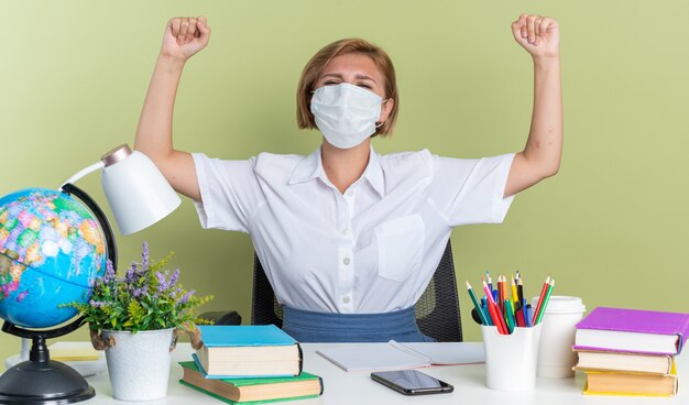 Gioiosa giovane studentessa bionda che indossa una maschera protettiva seduta alla scrivania con gli strumenti della scuola che guarda l'obbiettivo facendo sì gesto isolato sul muro verde oliva