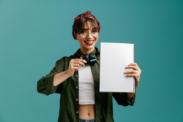 Gioiosa giovane studentessa che indossa occhiali bandana e cuffie intorno al collo che mostra un grande blocco note guardando la fotocamera che punta con la penna sul blocco note isolato su sfondo blu
