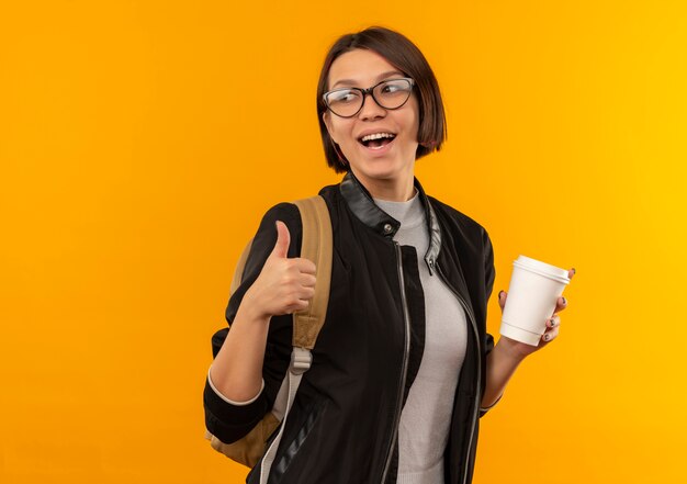 Gioiosa giovane studentessa con gli occhiali e borsa posteriore che tiene la tazza di caffè in plastica guardando il lato e mostrando il pollice in alto isolato sull'arancio