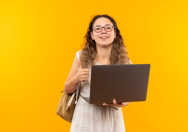 Gioiosa giovane studentessa graziosa con gli occhiali e borsa posteriore che tiene il computer portatile che mostra il pollice in su isolato su giallo