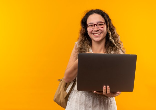 Gioiosa giovane studentessa graziosa con gli occhiali e borsa posteriore che tiene il computer portatile isolato su giallo