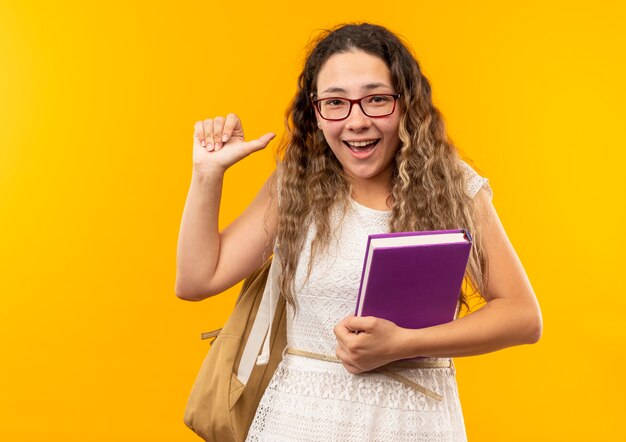 Gioiosa giovane studentessa graziosa con gli occhiali e borsa posteriore che tiene il libro che indica se stessa isolato su giallo