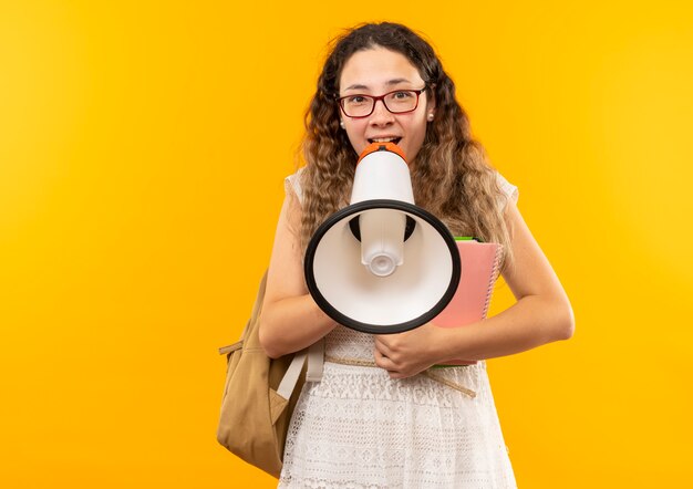 Gioiosa giovane studentessa graziosa con gli occhiali e borsa posteriore tenendo appunti parlando da altoparlante isolato su giallo