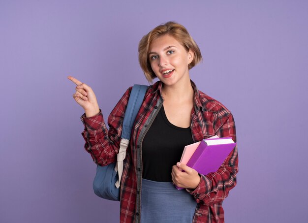 Gioiosa giovane studentessa slava che indossa uno zaino tiene i punti di libri e quaderni a lato