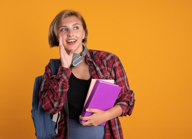 Gioiosa giovane studentessa slava con le cuffie che indossa lo zaino tiene la mano vicino alla bocca tiene libro e taccuino